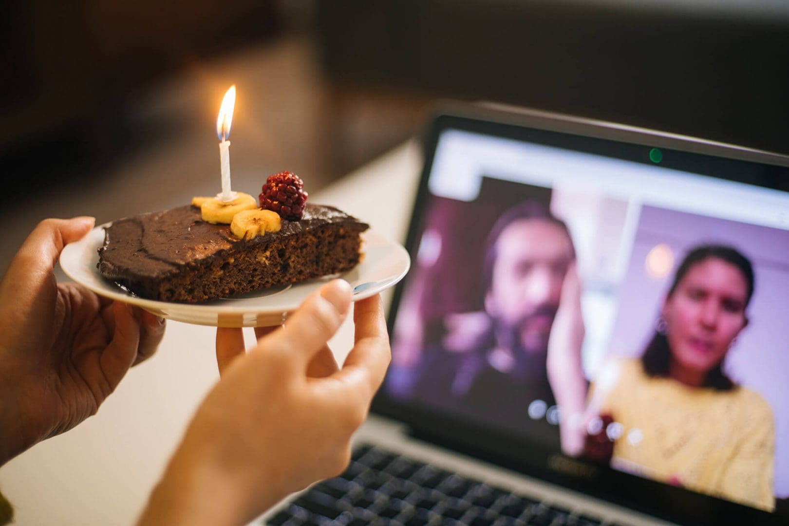 A person holding a cake with a candle in front of a laptop screen.